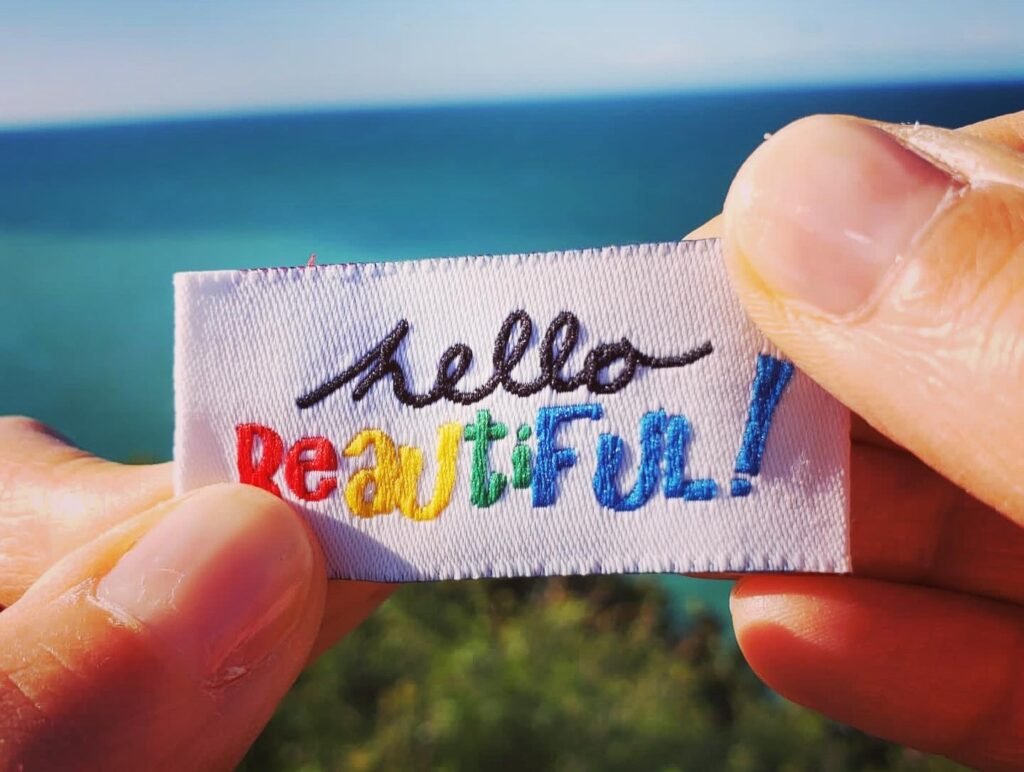 White hands hold up a colourful woven label reading, "Hello Beautiful" against a beautiful, blue sky and green bushes.